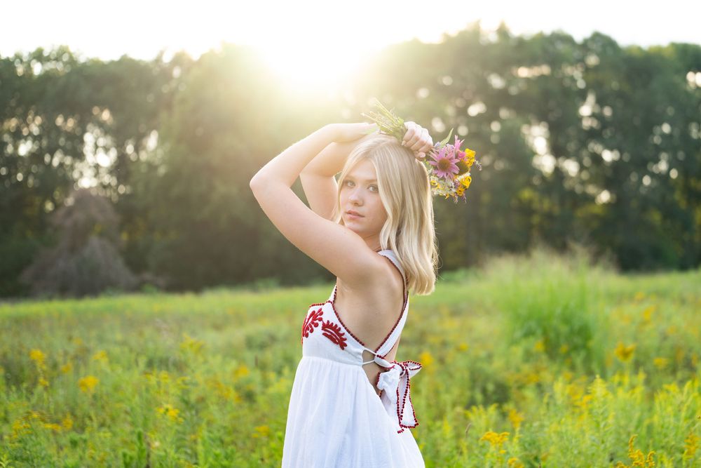 Blonde woman in white sundress with floral crown standing in sunny meadow with yellow wildflowers and trees.