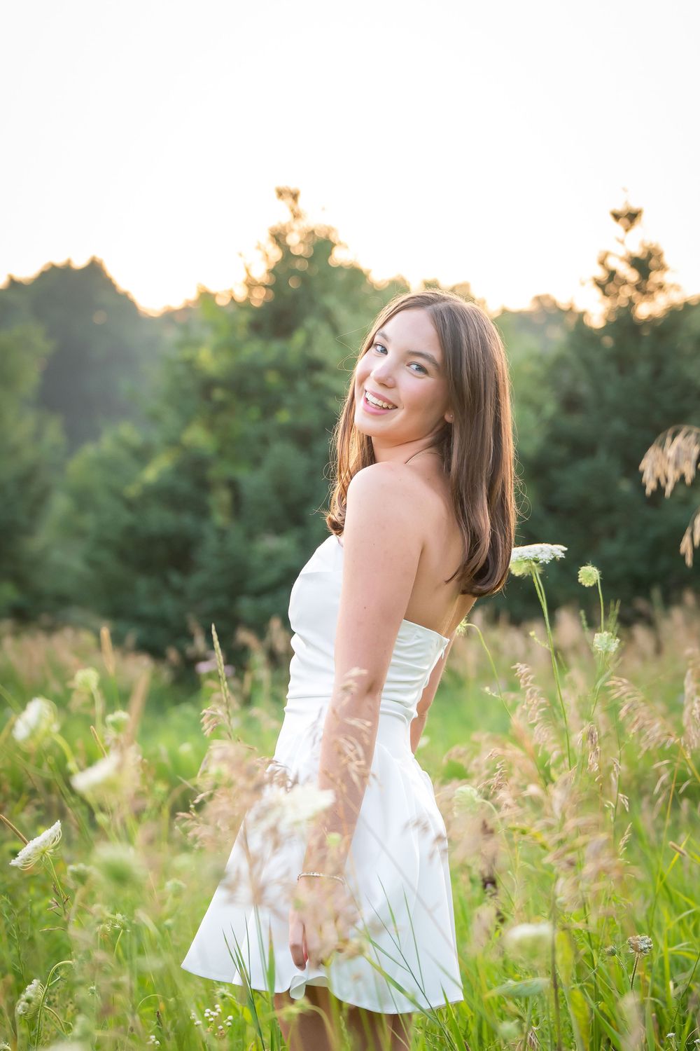 Young woman in white strapless dress smiling in a sunny wildflower meadow with green trees in background.