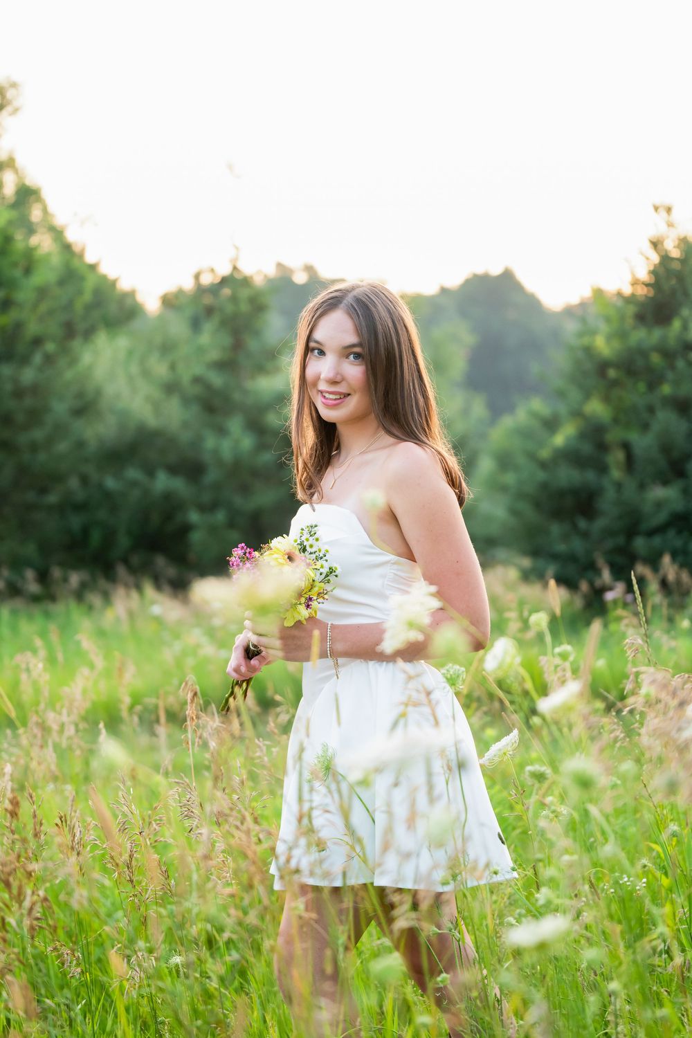 Young woman in white dress holding wildflowers smiling in a sunny green meadow with trees in the background.