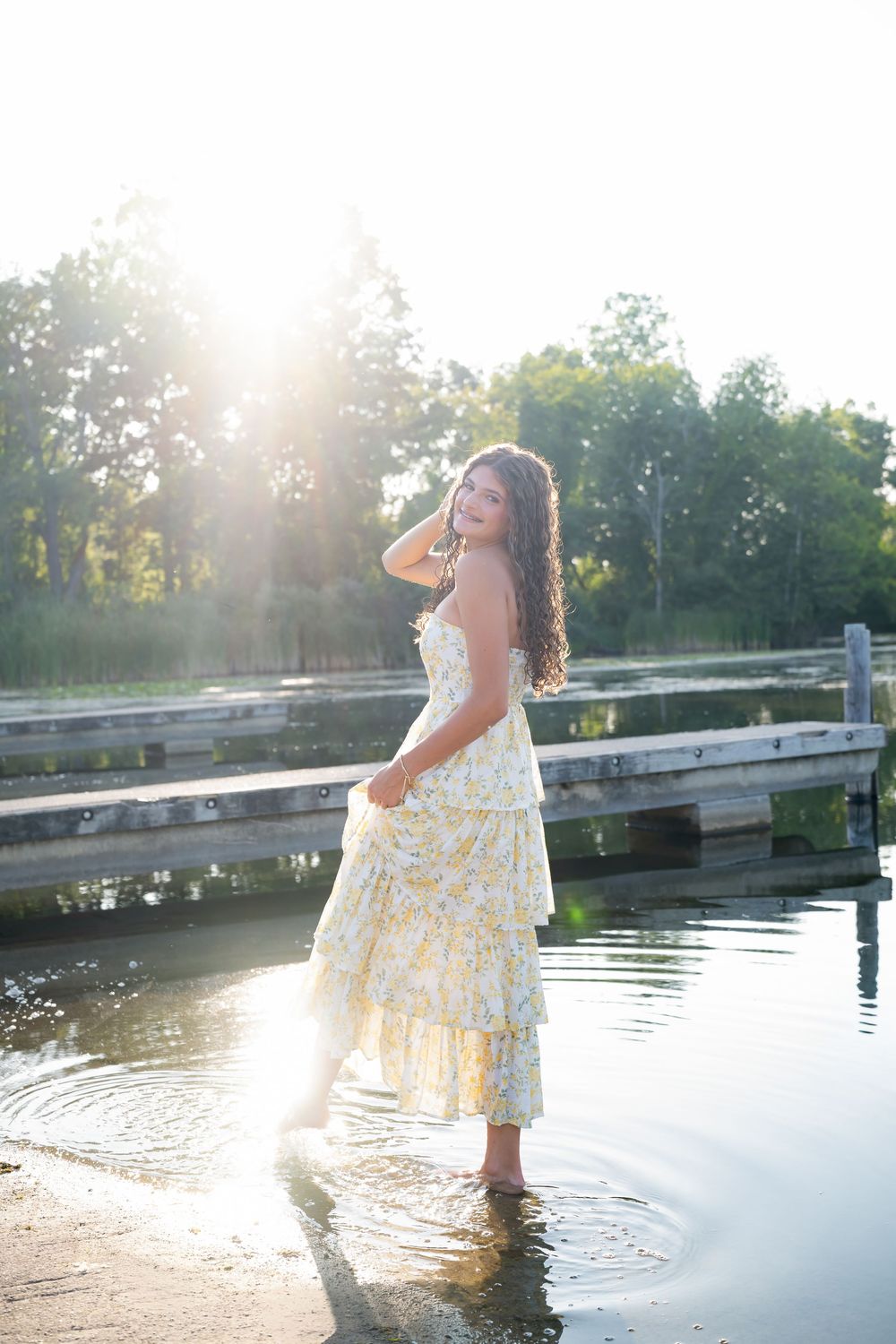 Young woman in floral sundress wading in shallow water near a dock, backlit by golden sunset light.