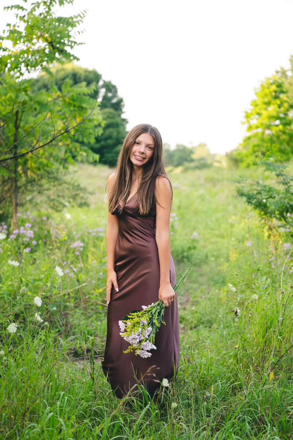 Young woman in brown maxi dress holding wildflowers, smiling in a lush green meadow on a sunny day.