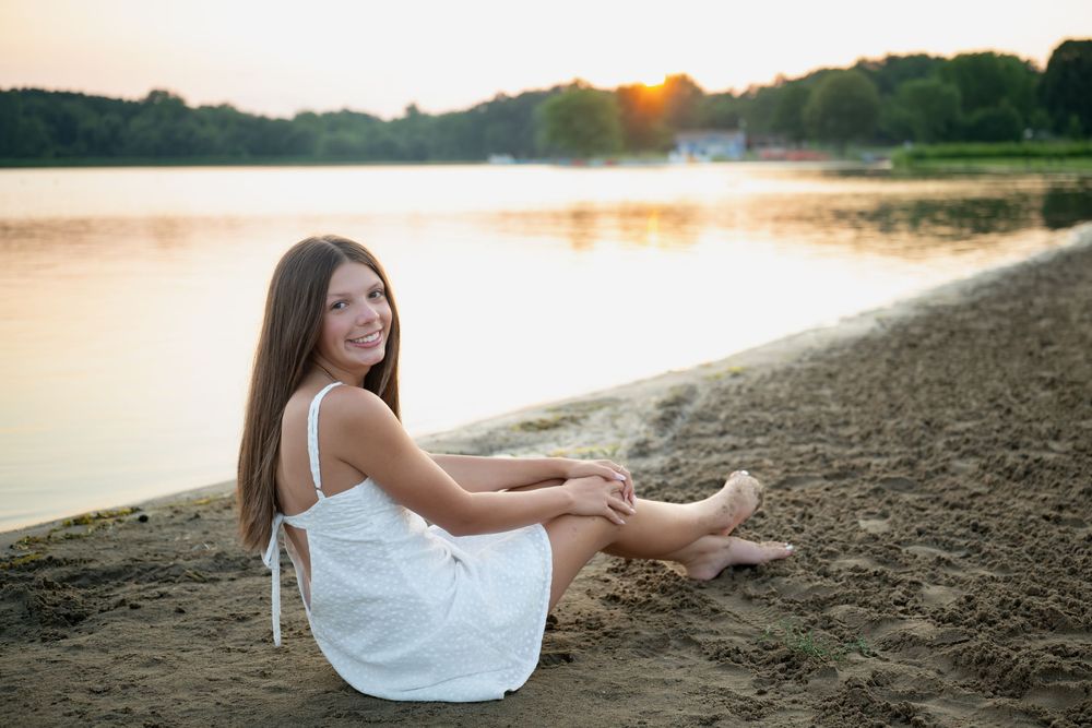 A smiling young woman in a white dress sits barefoot on a sandy beach shore at golden sunset.