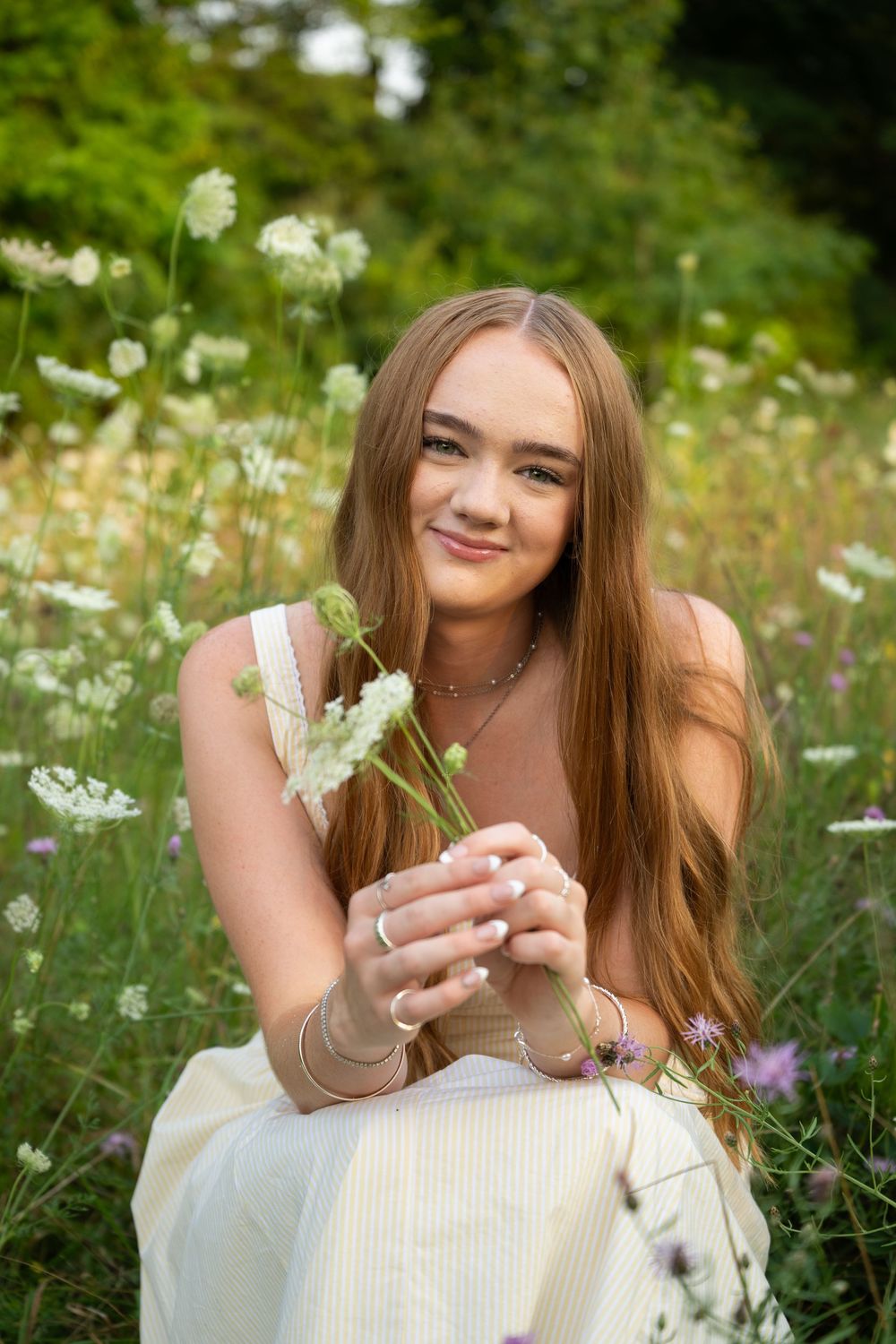 Young woman with long auburn hair sitting in a wildflower meadow, smiling and holding white flowers.