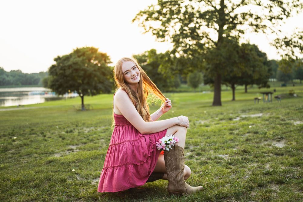 Smiling redhead woman in pink dress and cowboy boots sitting on grass in a sunny park with trees and lake.