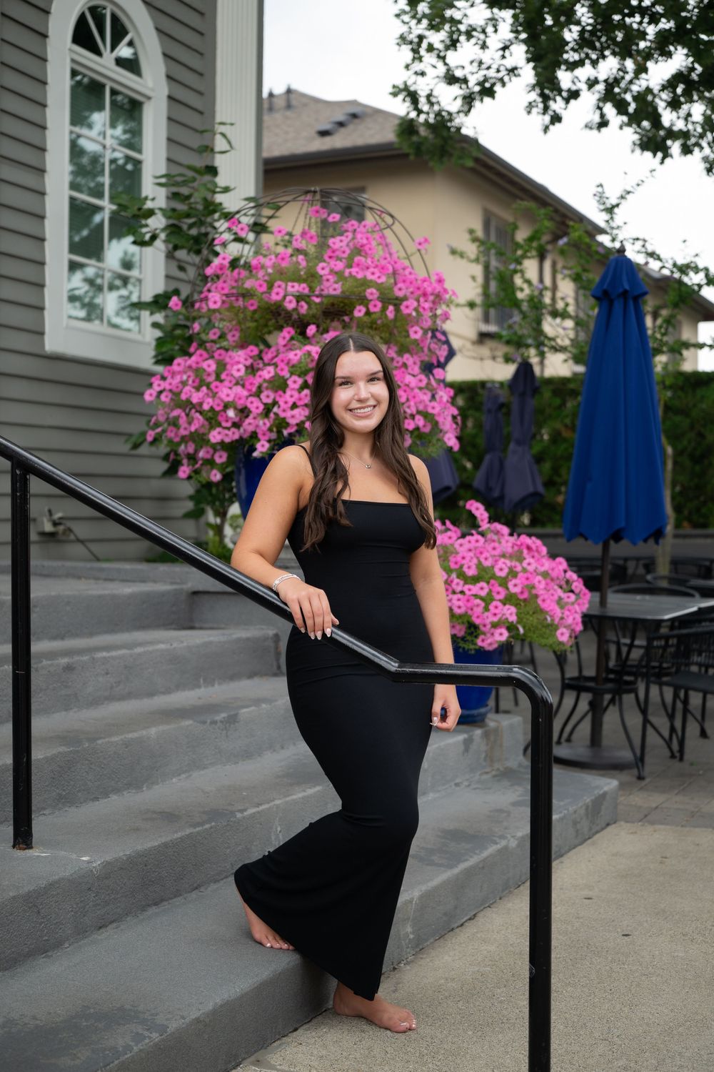 Young woman in black jumpsuit poses barefoot on steps with vibrant pink hanging flowers in outdoor setting.
