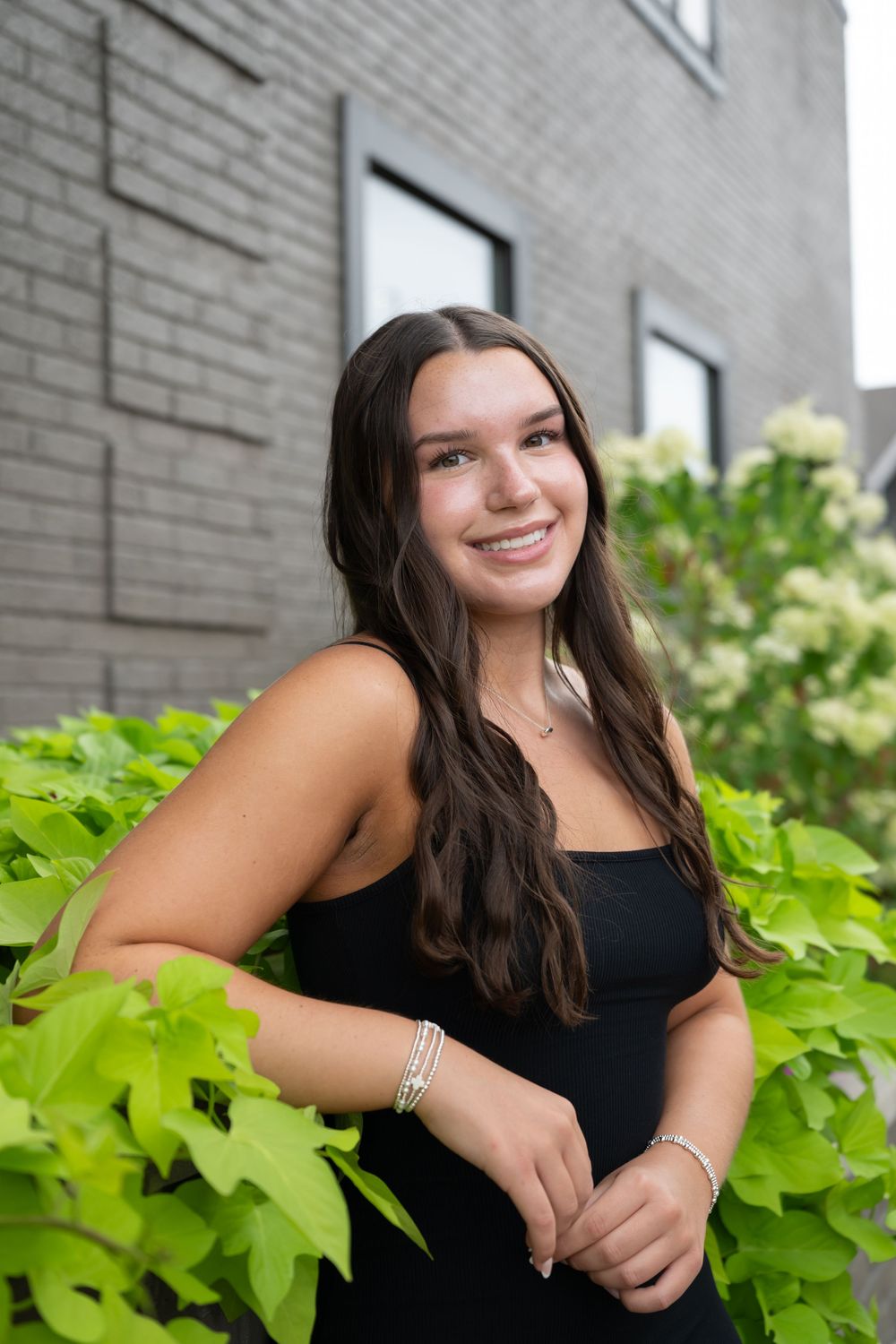 Young woman with long dark hair smiling outdoors in a black dress, surrounded by lush green plants near a brick building.