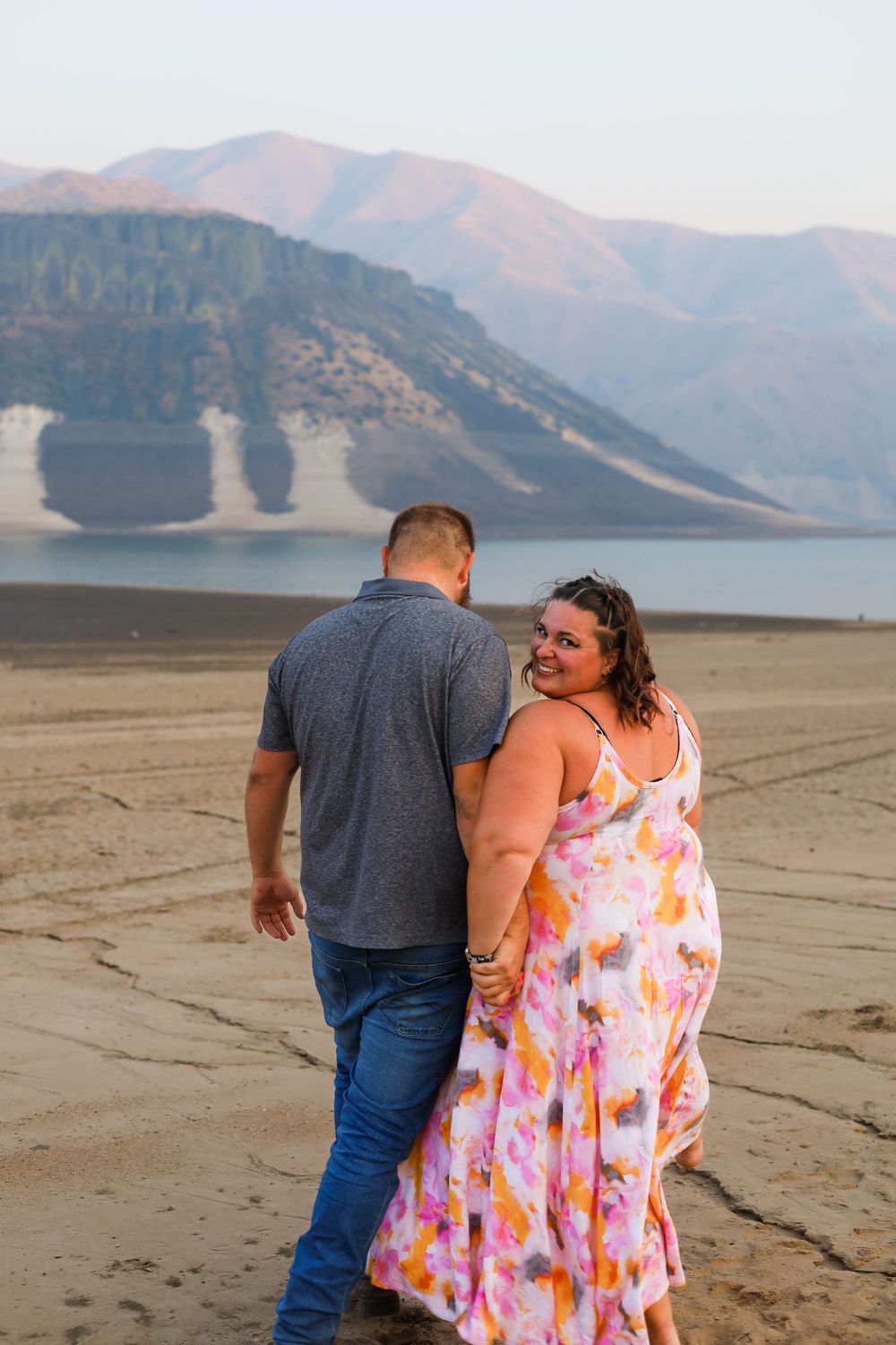 A couple walking along a scenic beach with mountains and lake in the background.