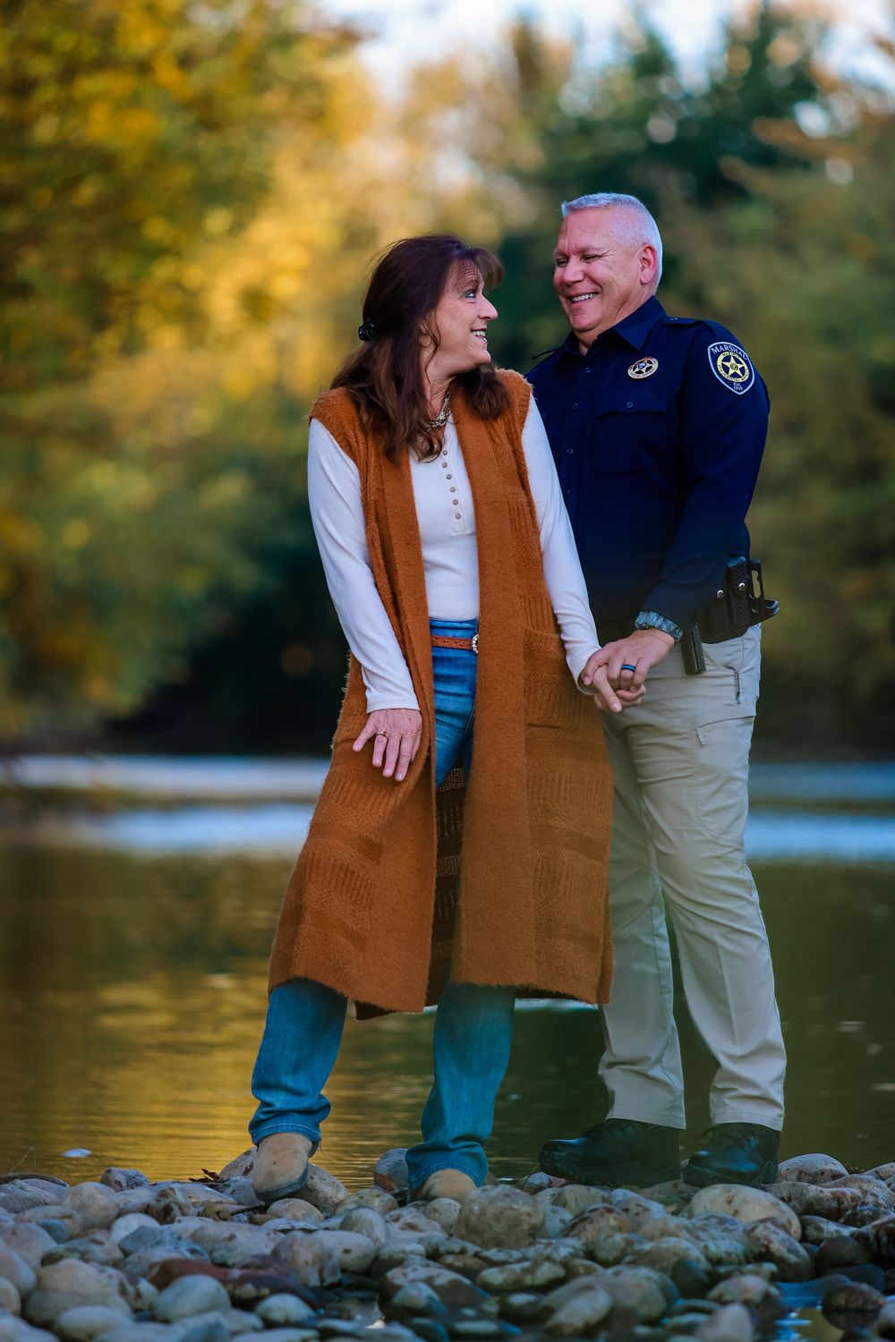 A couple stands together on rocky riverbank during golden hour in autumn.