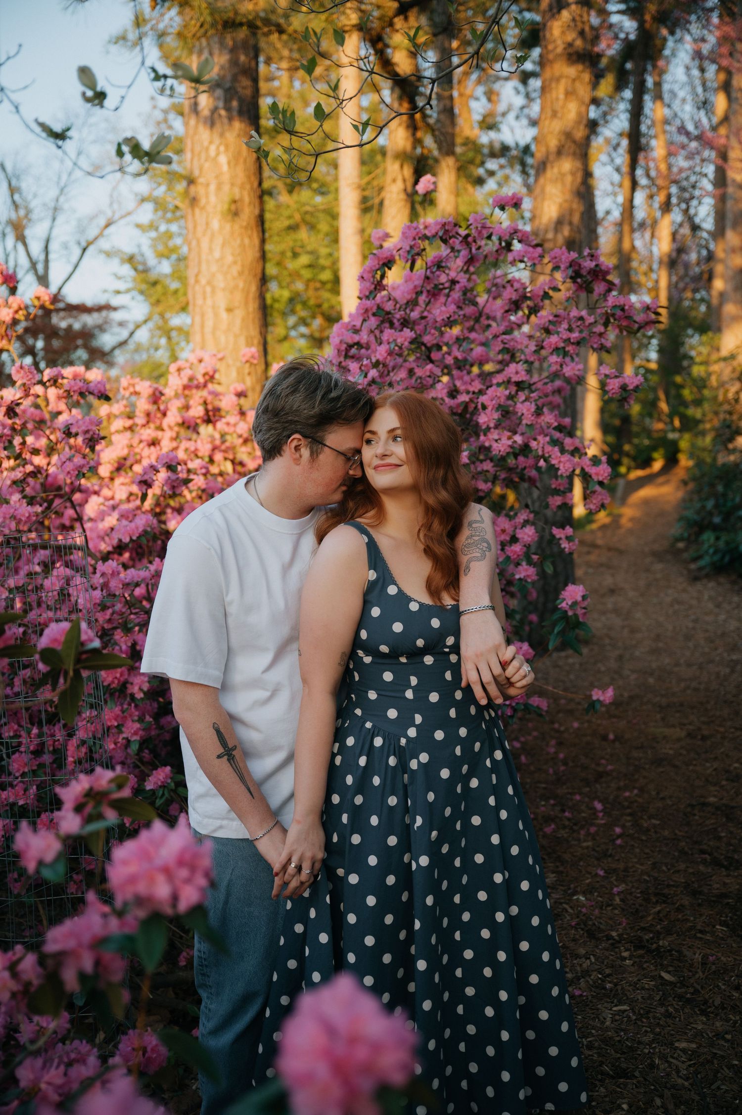 Couple embracing among pink blooming rhododendrons at golden hour, woman in polka dot dress, man in white tee.