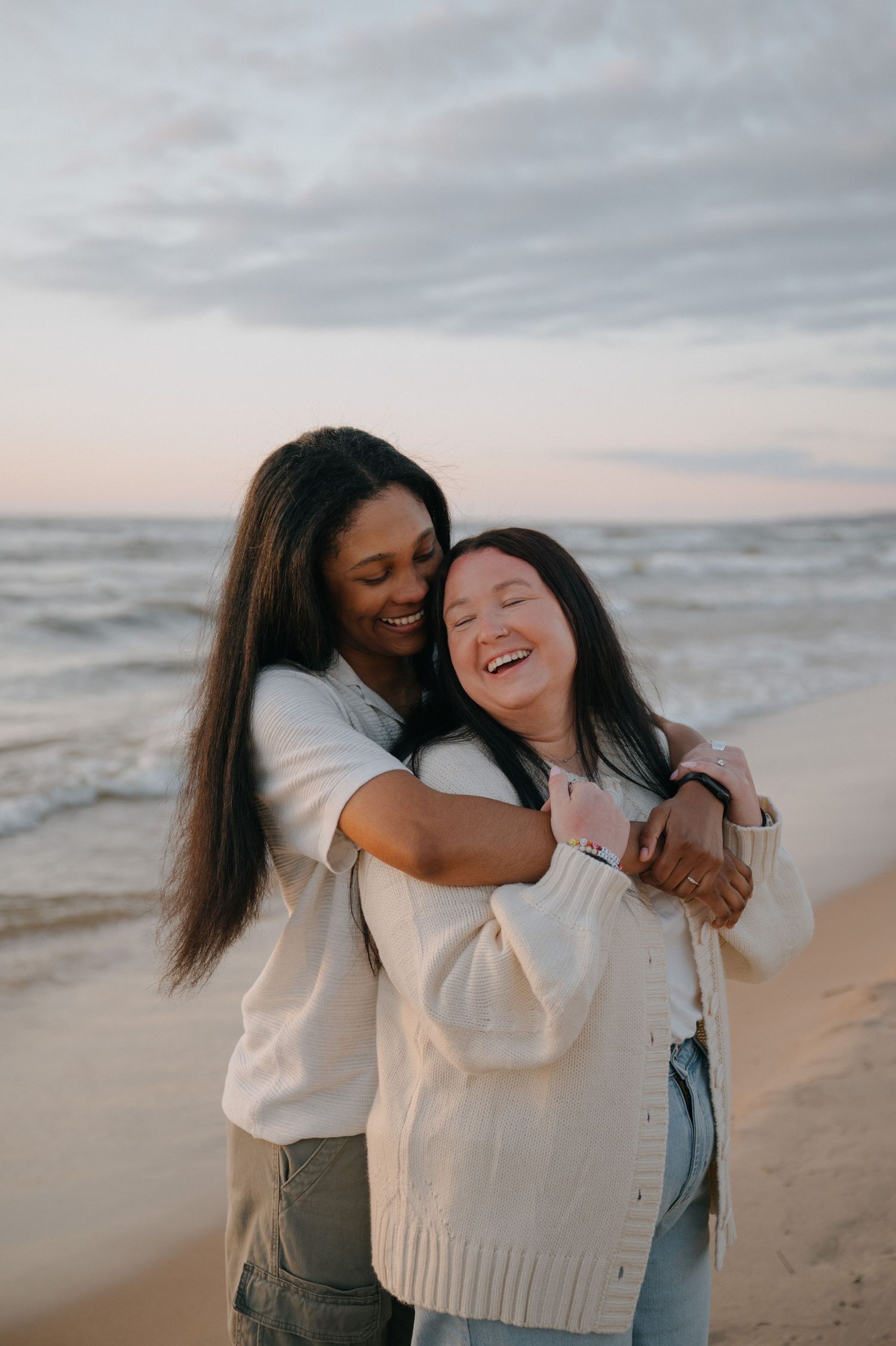 Two women sharing a joyful hug on a sandy beach at sunset, laughing and embracing warmly.