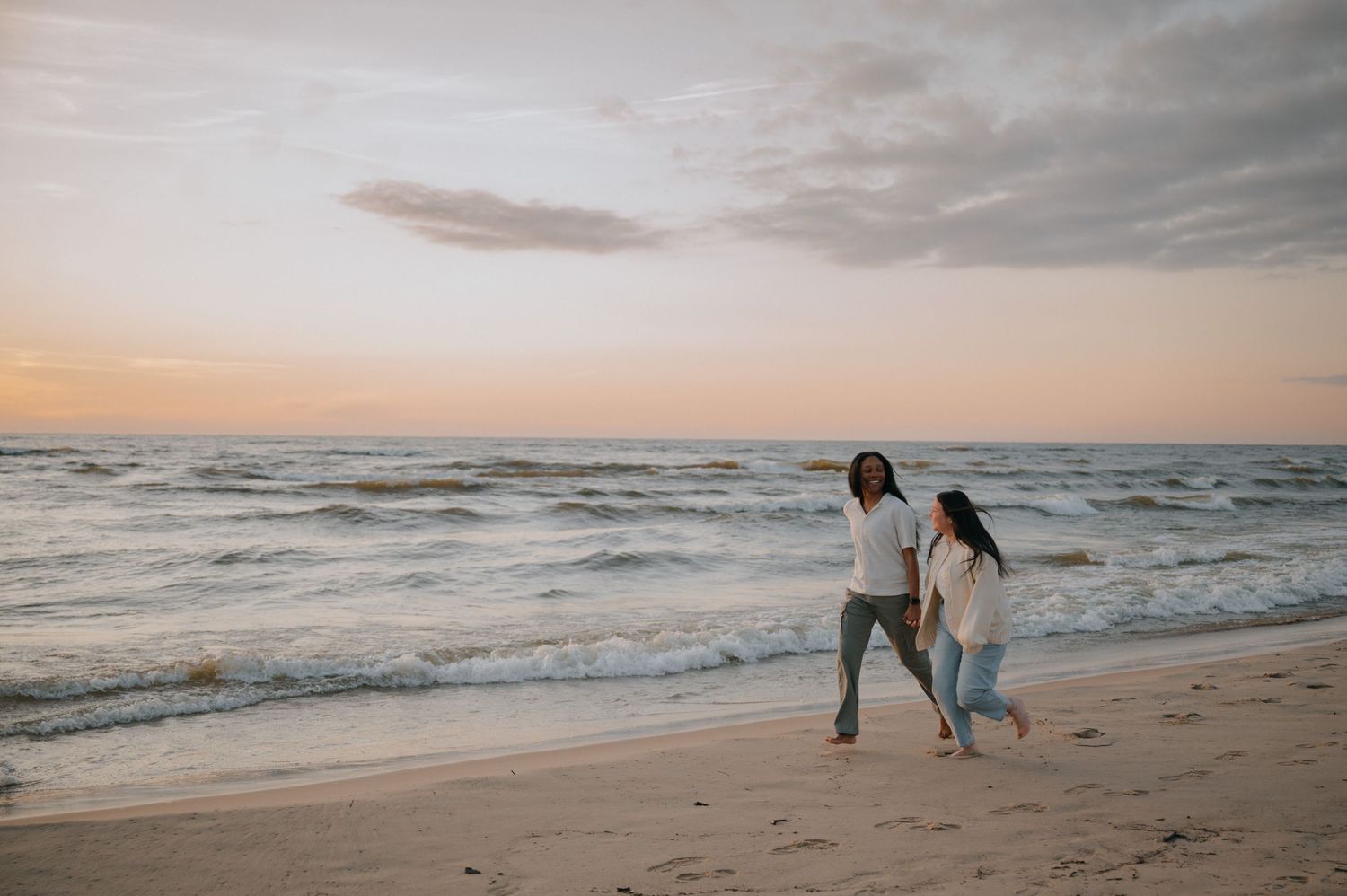 Two women walk along a sandy beach at sunset, waves crashing behind them under a pastel sky.