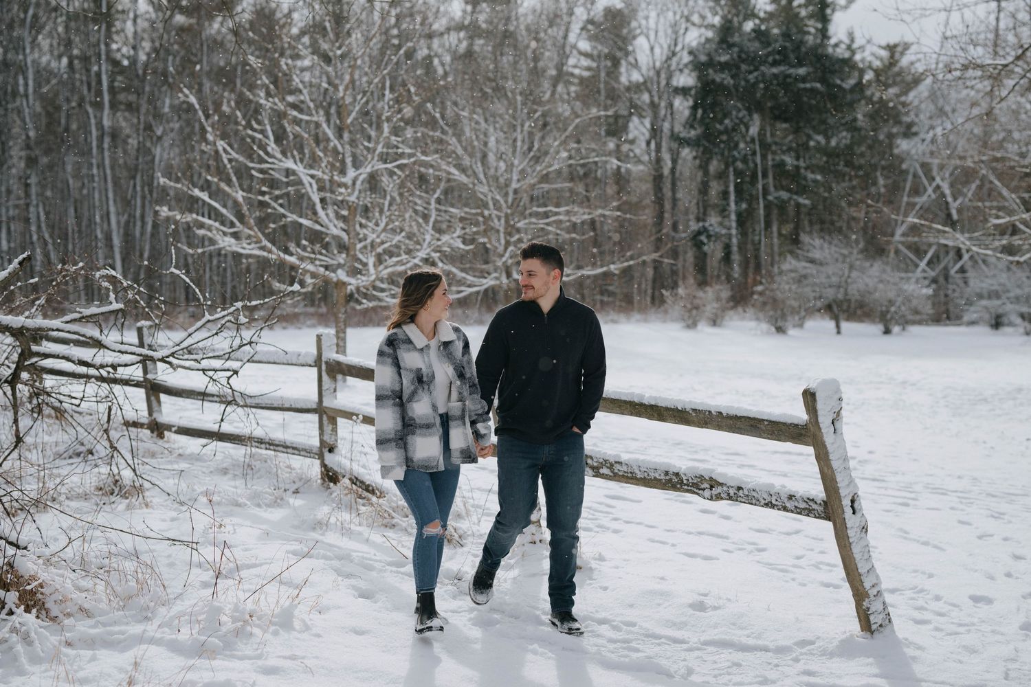 A couple takes a romantic winter walk along a snow-covered wooden fence with bare trees in the background.