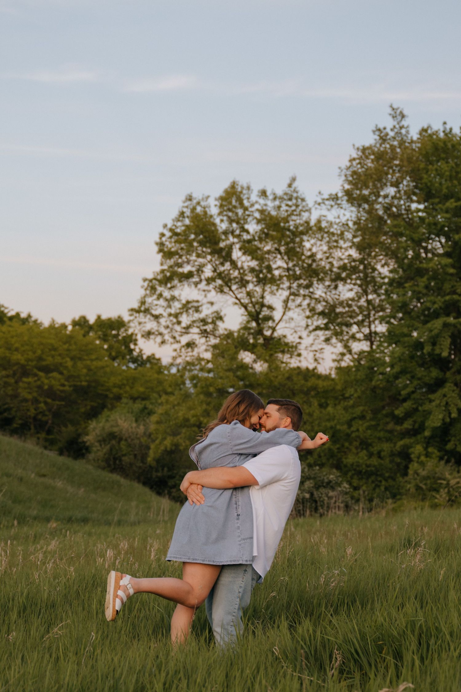 A couple embraces in a sunlit meadow surrounded by green trees during golden hour.
