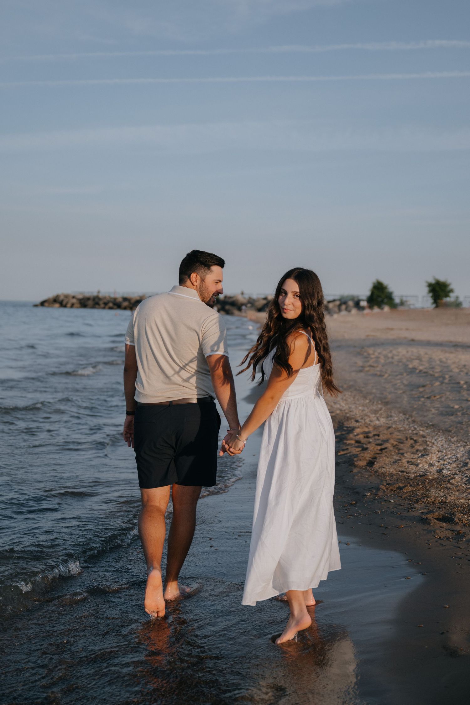 A couple holding hands while walking barefoot along the beach at sunset with waves crashing beside them.