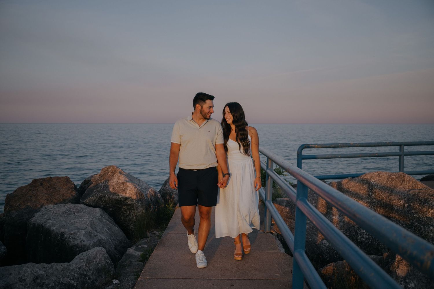 A couple walks together at sunset on a wooden pier overlooking the ocean and rocky shoreline.