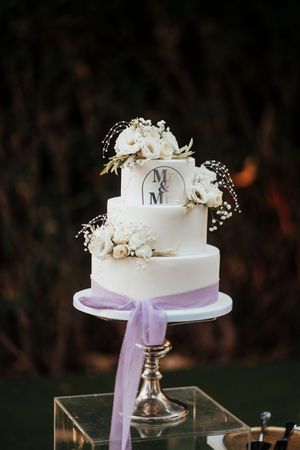 Elegant white wedding cake with purple ribbon accent displayed on silver stand against dark background.
