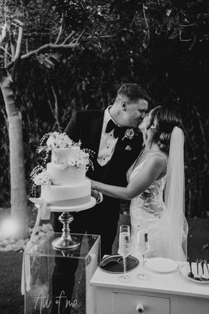 A black and white wedding photo of a couple sharing an intimate moment while cutting their elegant tiered cake outdoors.