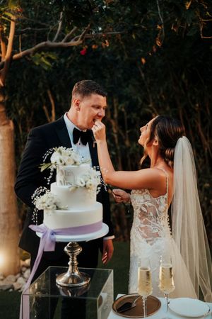 Wedding cake with purple ribbon displayed on silver stand at outdoor reception as couple shares sweet moment.