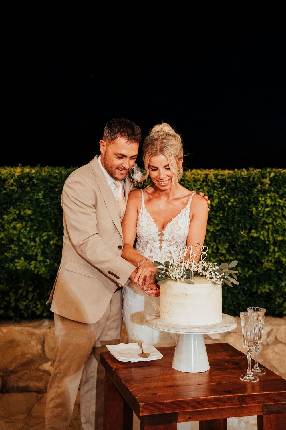 A couple cuts their elegant white wedding cake together at an outdoor reception during golden hour.