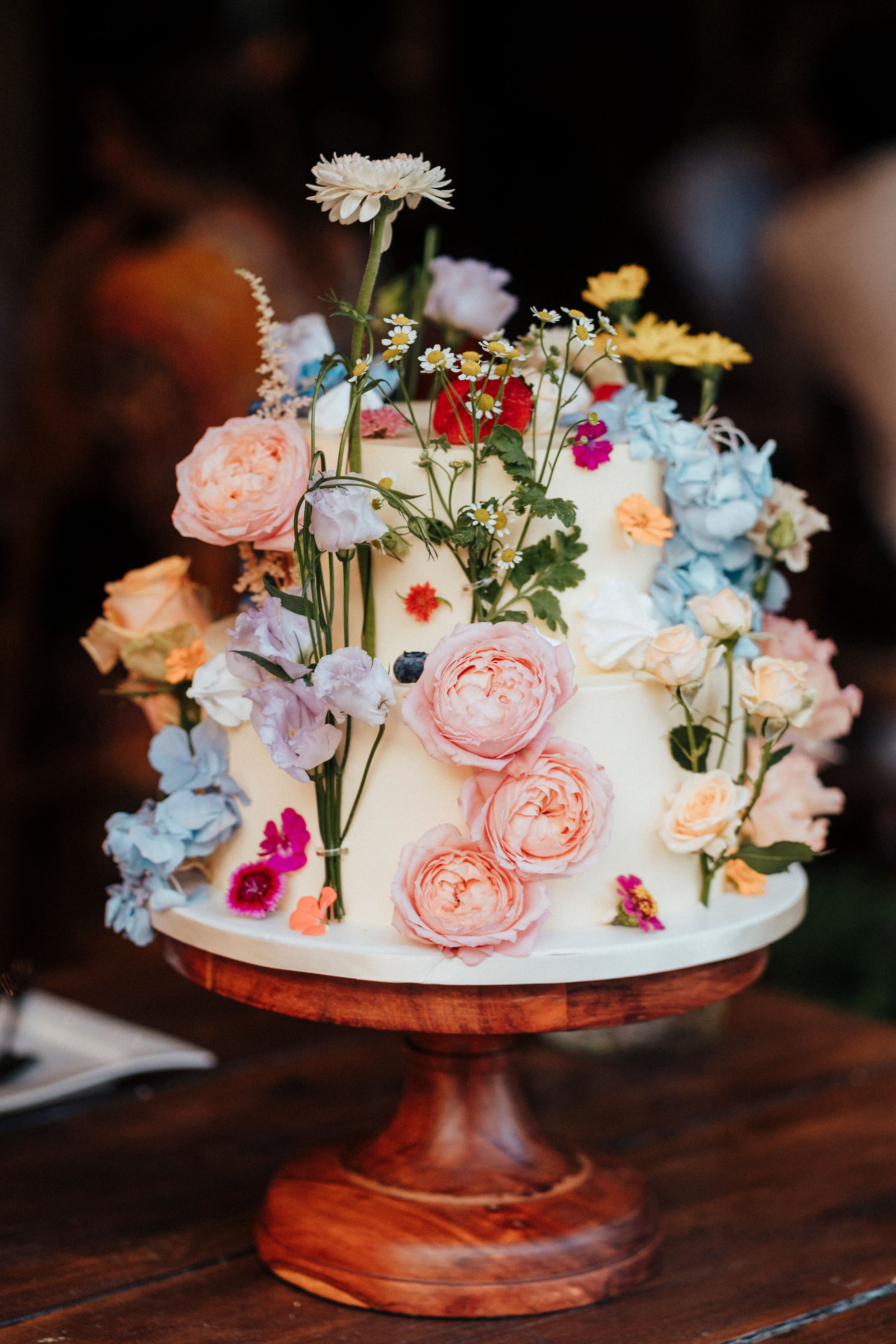 A white wedding cake decorated with fresh pastel flowers including roses, hydrangeas and wildflowers on a wooden cake stand.