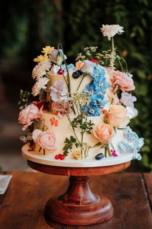 A rustic wooden cake stand displays a delicate floral arrangement with peach roses, blue hydrangeas and white blooms.