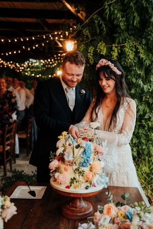A couple cuts their elegant three-tiered wedding cake adorned with pastel flowers under romantic string lights.