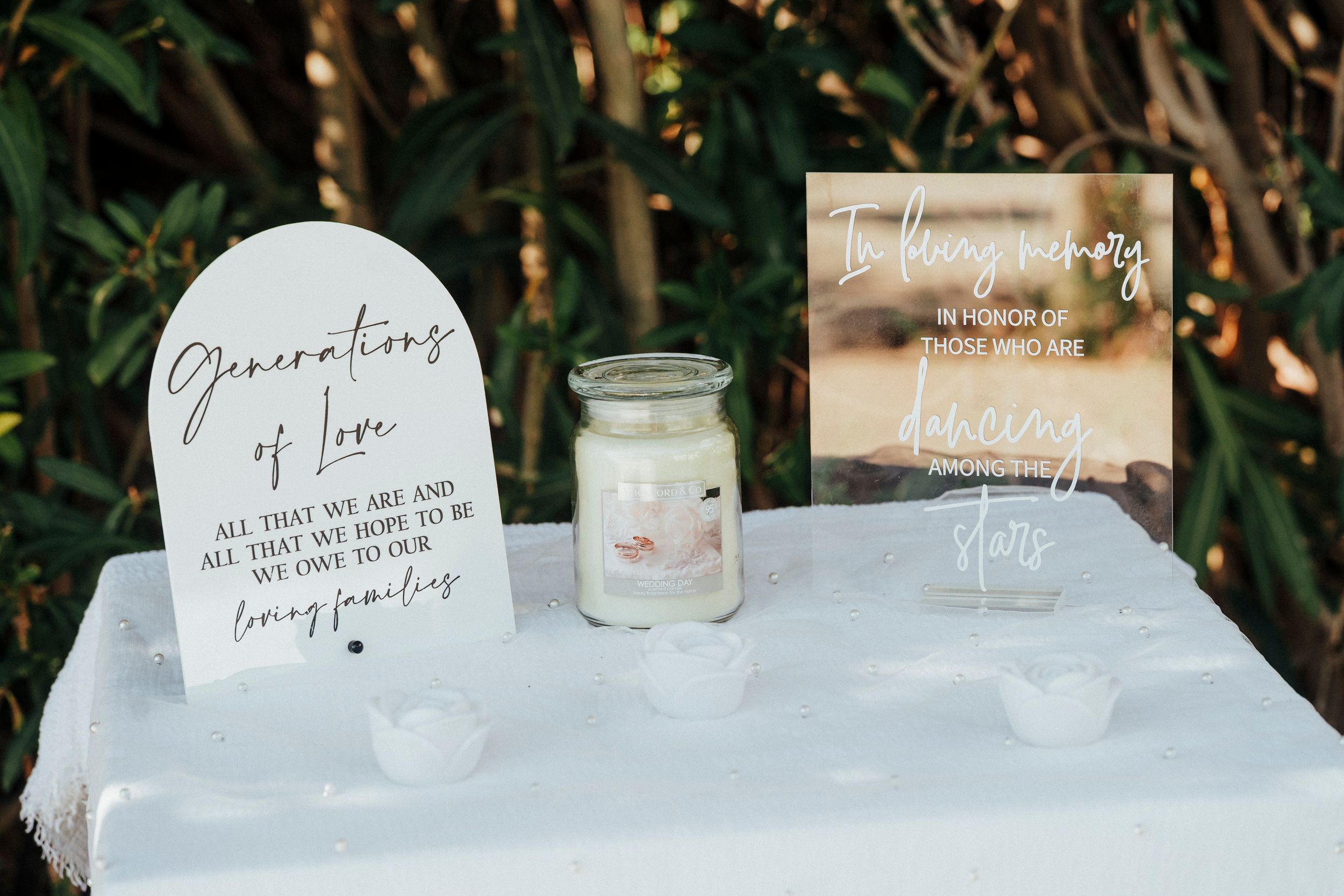 Wedding memorial signs and candle displayed on white table with greenery backdrop for remembrance of loved ones.