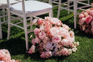 Pink and peach floral arrangements with roses and mixed blooms rest on grass beside white chairs at an outdoor event.