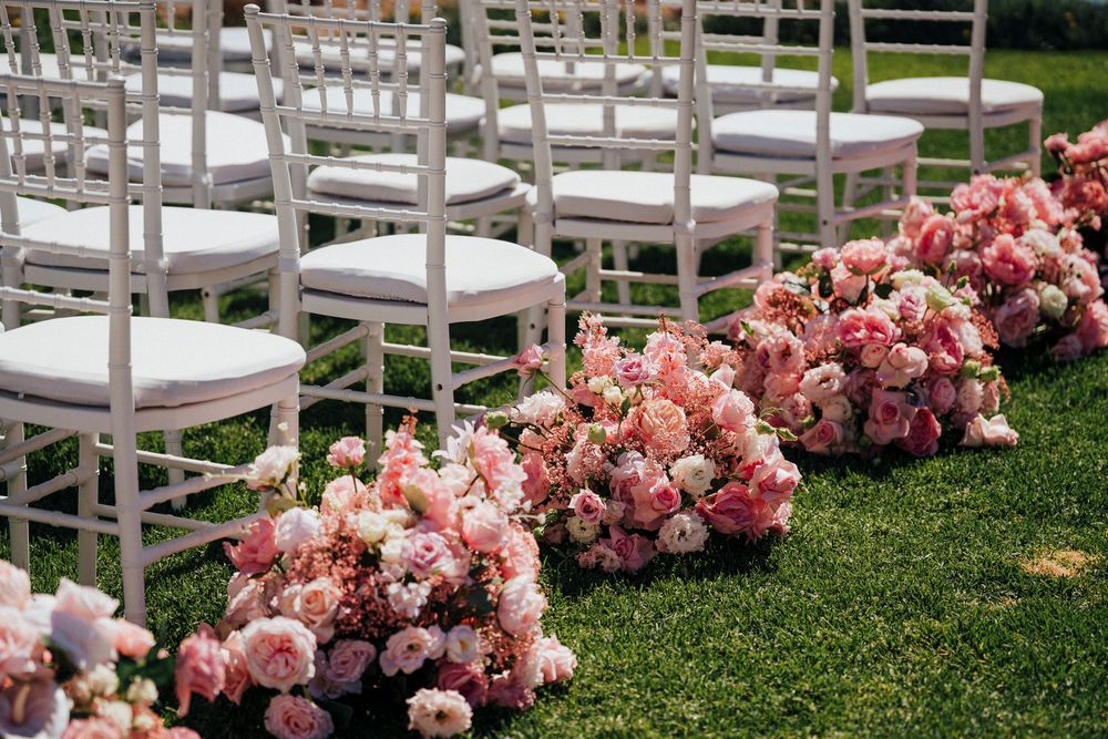 Pink floral arrangements line the aisle between white chairs on a lush green lawn at an outdoor wedding ceremony.