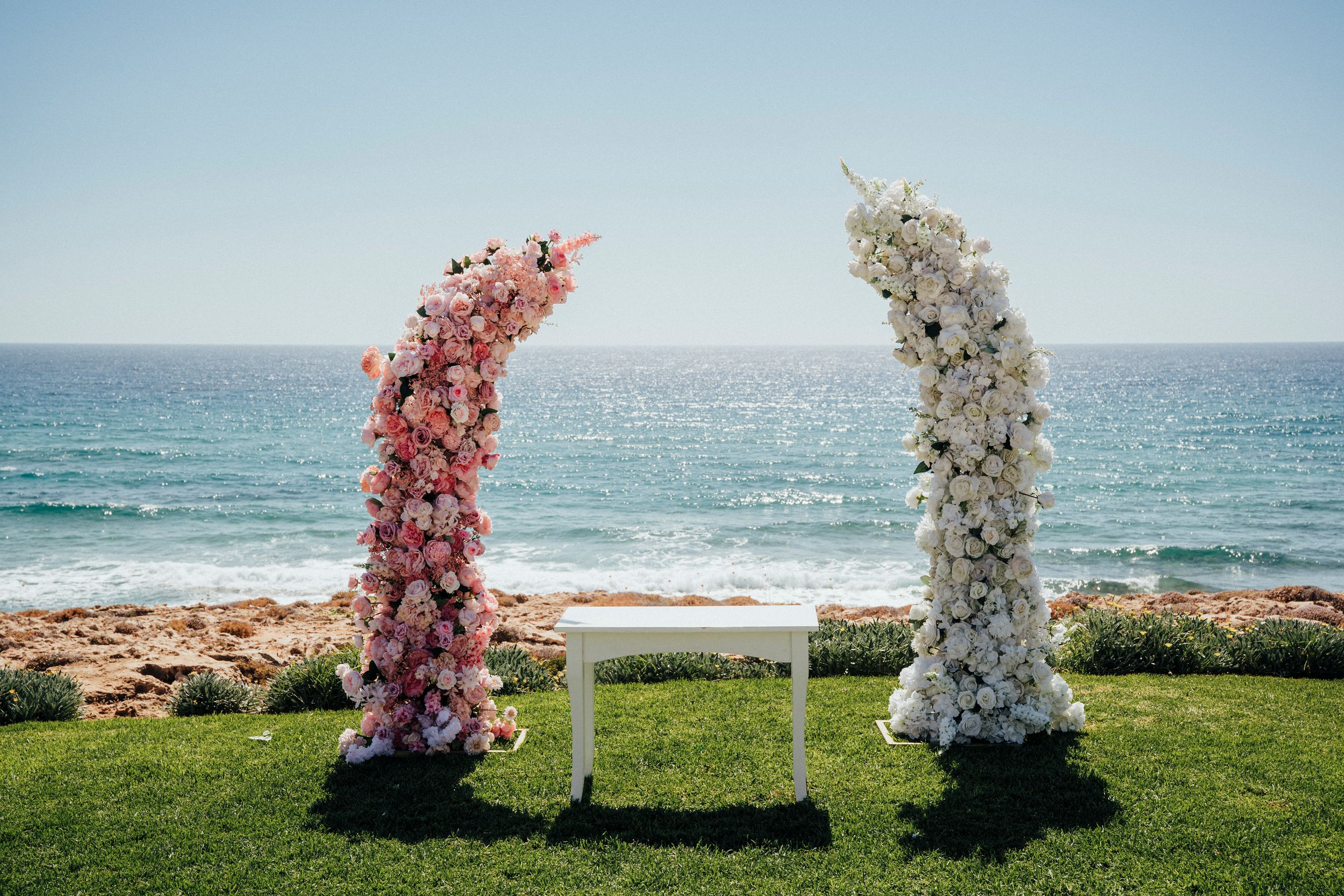 Pink and white floral wedding arches stand against a scenic ocean backdrop on a grassy cliff overlooking the sea.
