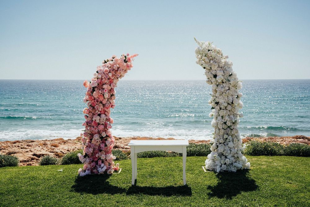 Pink and white floral wedding arches stand against a scenic ocean backdrop on a grassy cliff overlooking the sea.