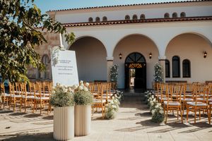 Rustic wooden chairs line an elegant Spanish-style courtyard with white stucco walls and arched walkways.