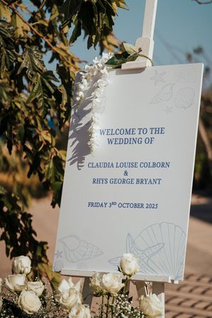 Wedding welcome signs with white flowers and blue text displayed outdoors against green foliage background.