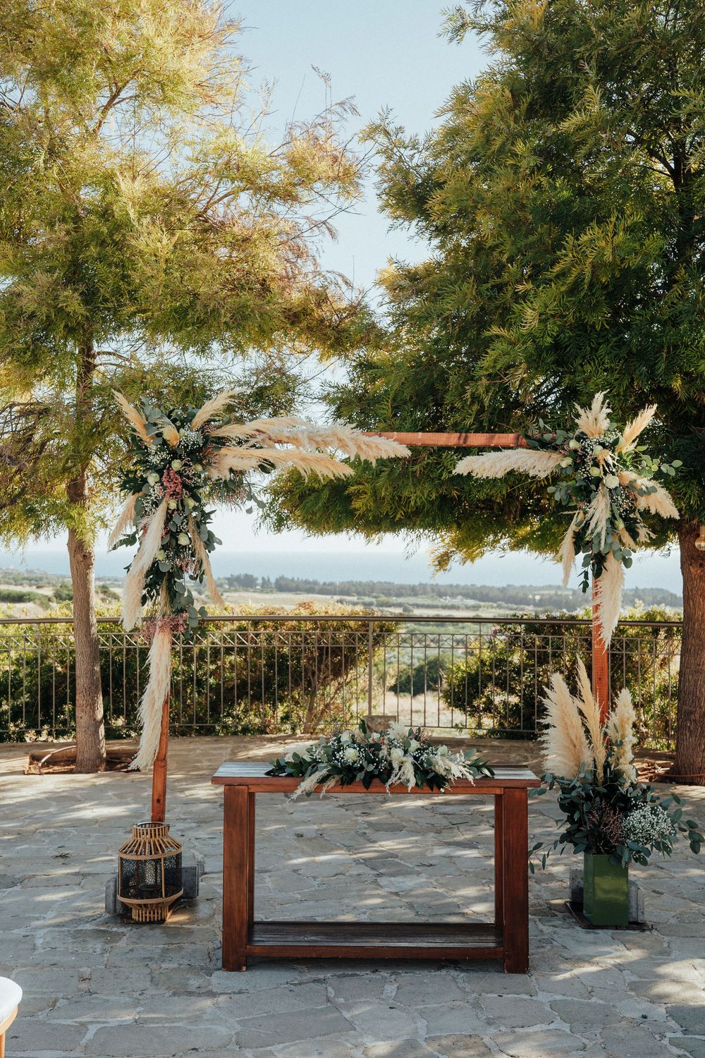 Outdoor wedding ceremony setup with wooden benches and floral arrangements under palm trees at a scenic overlook.