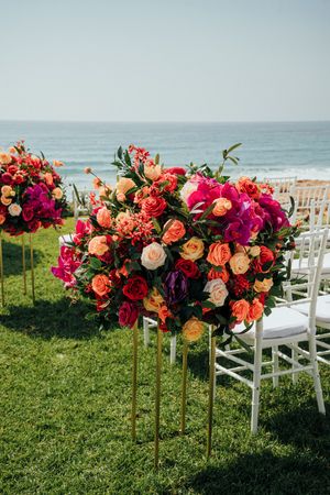 Vibrant coral and magenta floral arrangements line a white chair aisle at an oceanfront wedding ceremony setup.