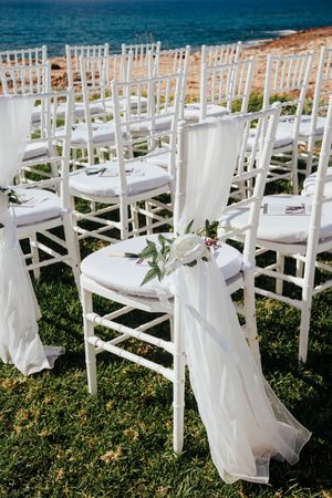 White wooden chairs with delicate fabric decorations arranged in rows for an outdoor wedding ceremony by the ocean.