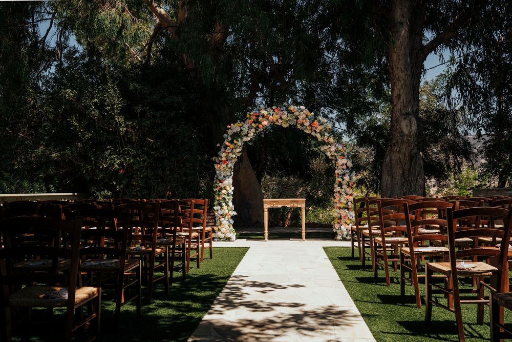 Outdoor wedding ceremony setup with wooden chairs and floral arch on green lawn under trees.