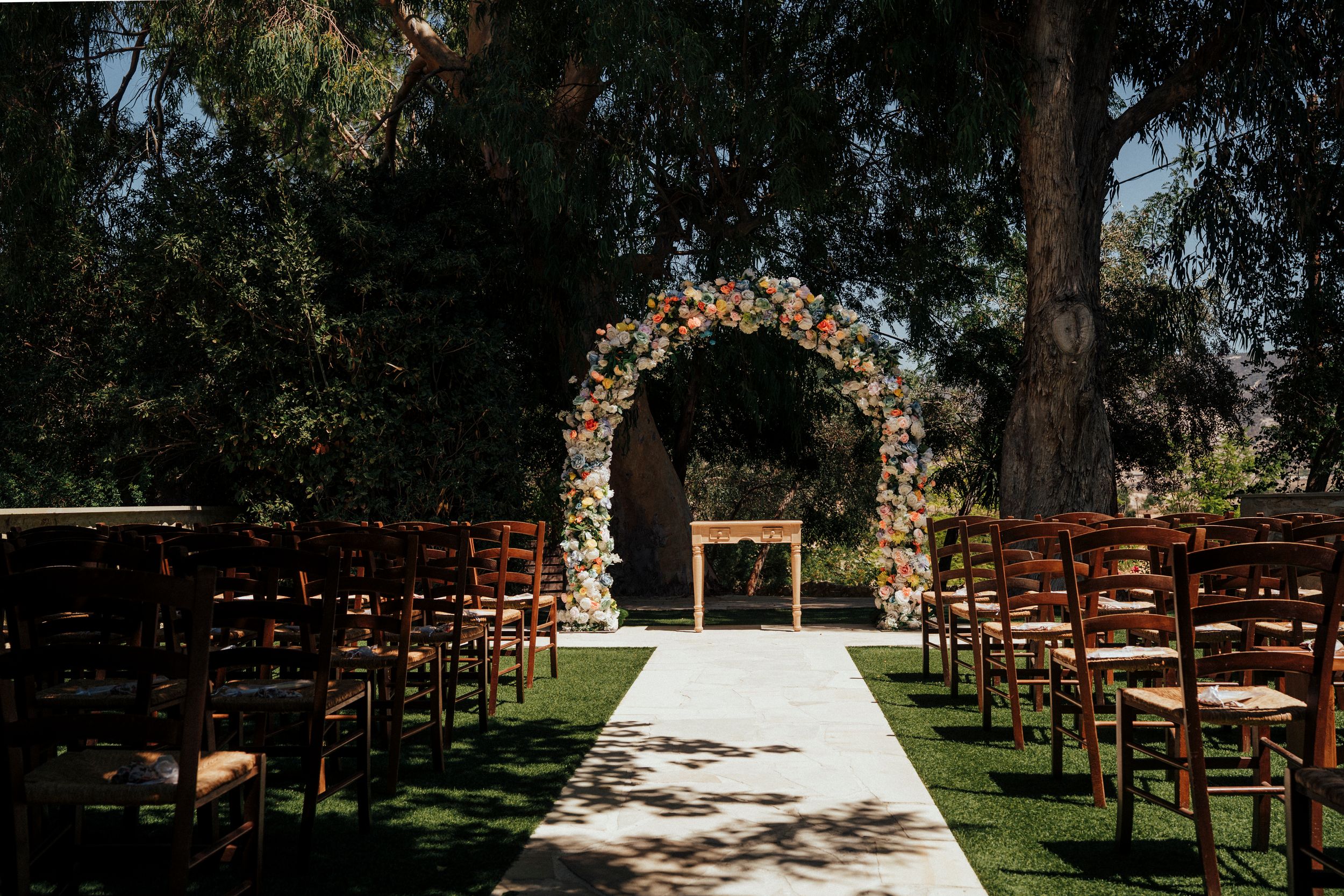 Outdoor wedding ceremony setup with wooden chairs and floral arch on green lawn under trees.
