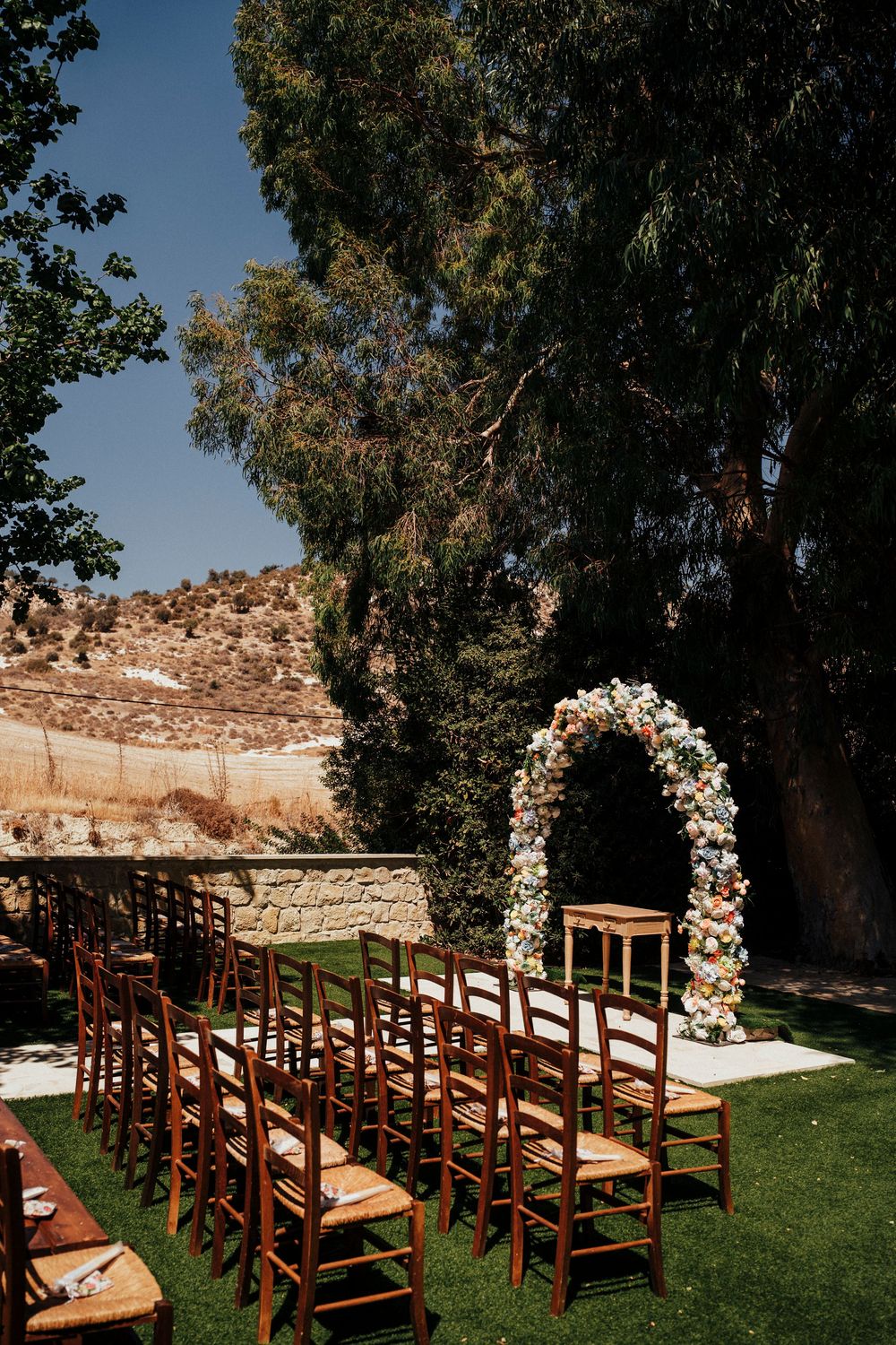 Wooden chairs arranged for an outdoor wedding ceremony with a floral arch and eucalyptus trees in the background.