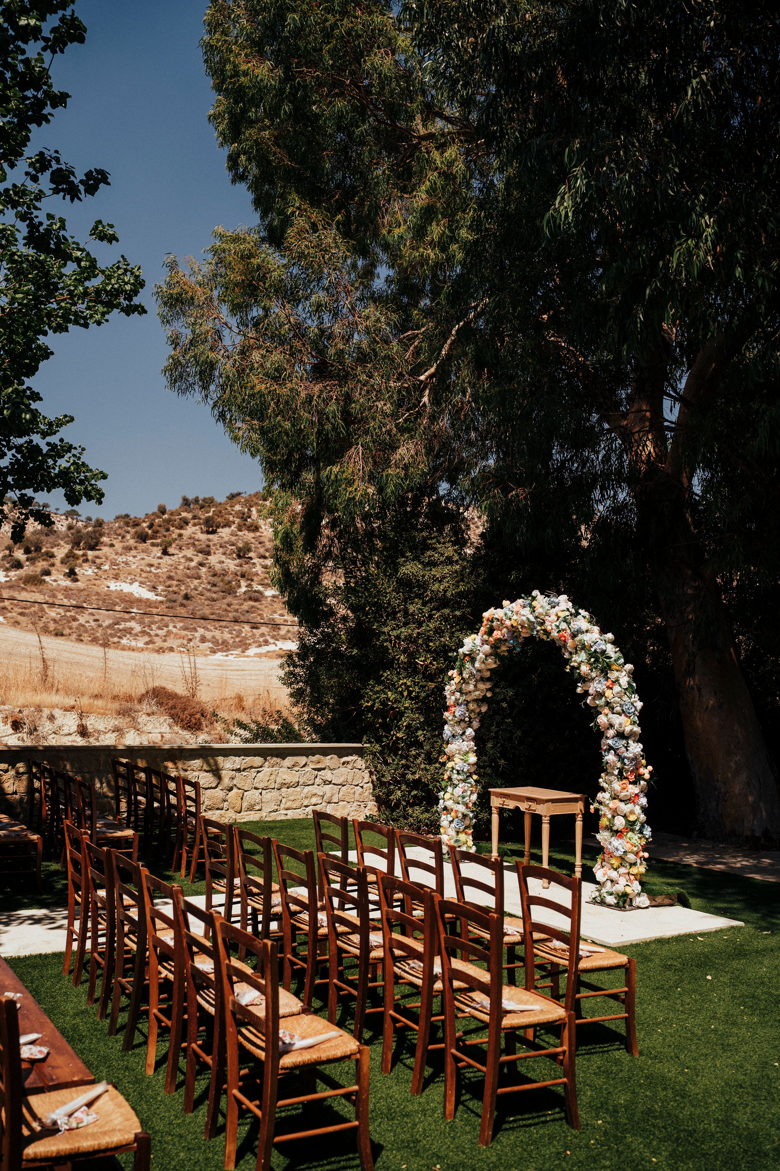 Wooden chairs arranged for an outdoor wedding ceremony with a floral arch and eucalyptus trees in the background.