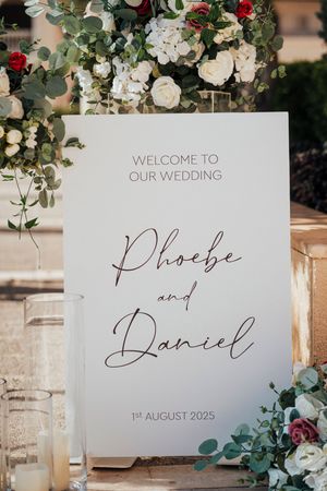 Wedding welcome sign decorated with eucalyptus leaves and roses features elegant calligraphy text on white paper.