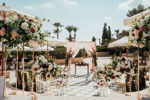 Outdoor wedding ceremony setup with floral arches, wooden chairs and palm trees against a Mediterranean backdrop.