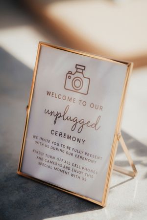 Gold-framed sign at wedding ceremony requesting guests to turn off phones for an unplugged celebration.