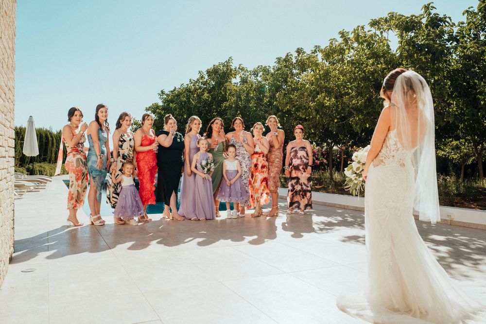 A wedding party stands together in a line outdoors with trees and greenery in the background.