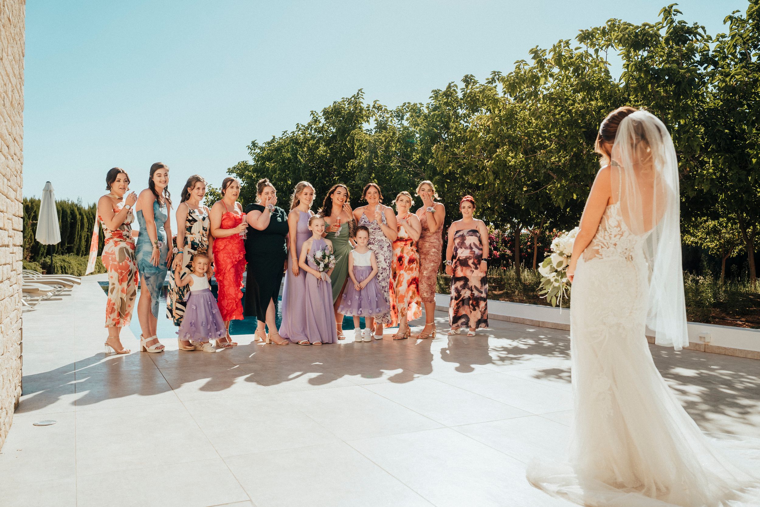 A wedding party stands together in a line outdoors with trees and greenery in the background.