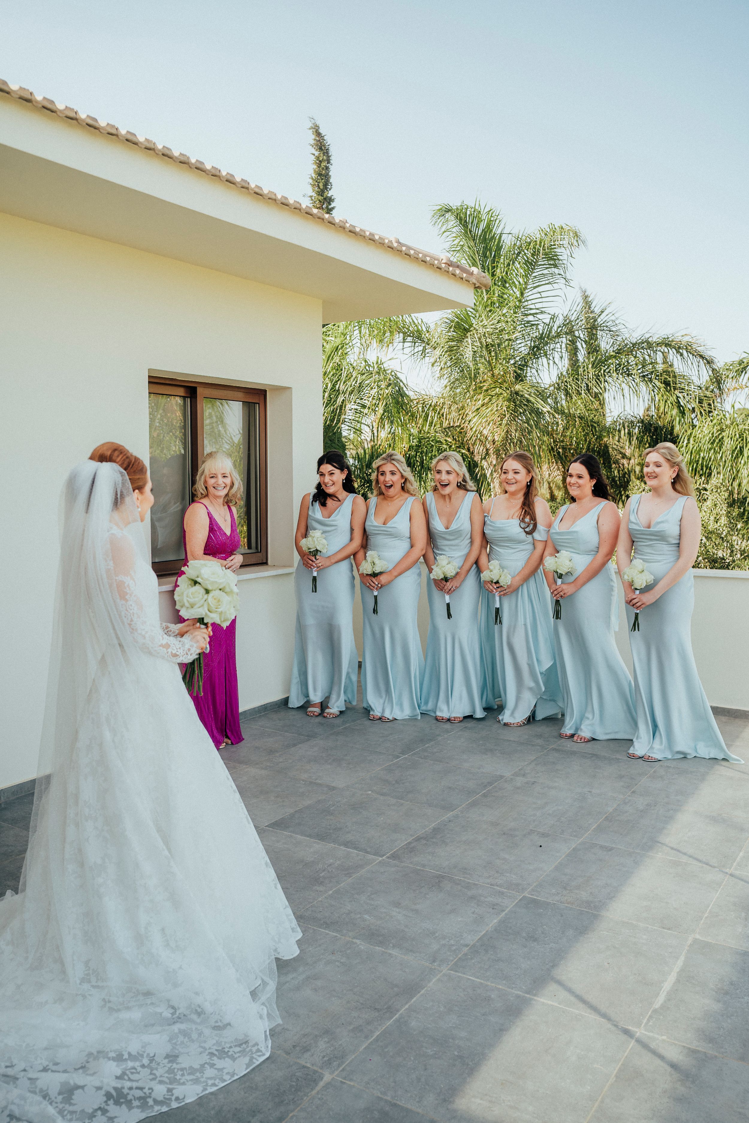 A group in pale blue bridesmaid dresses and a person in white stand together on a sunny outdoor terrace with palm trees.