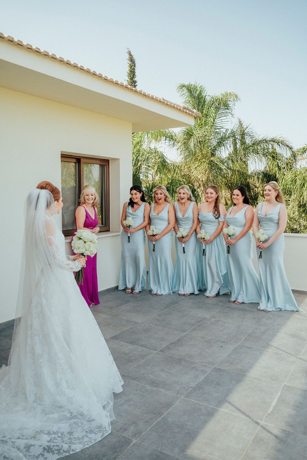 A group in pale blue bridesmaid dresses and a person in white stand together on a sunny outdoor terrace with palm trees.
