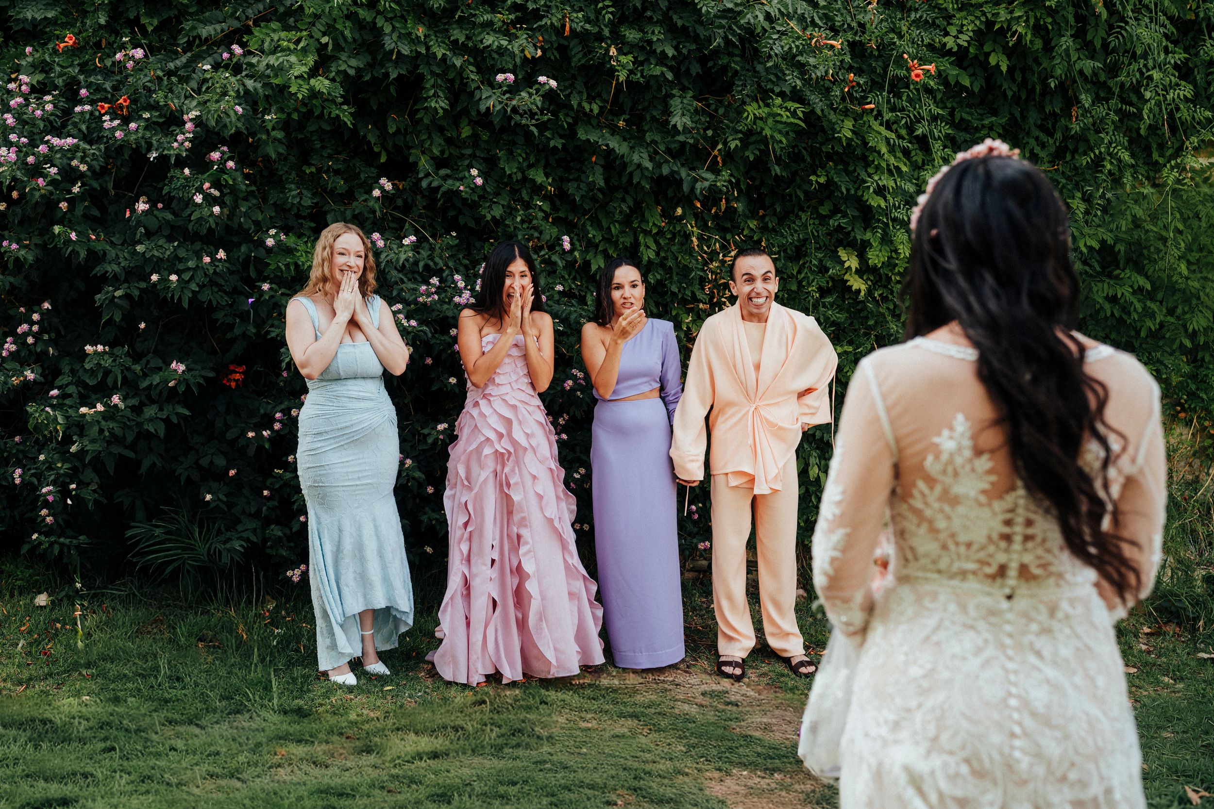 Wedding party members in pastel dresses and suits pose together in a garden setting during a summer celebration.