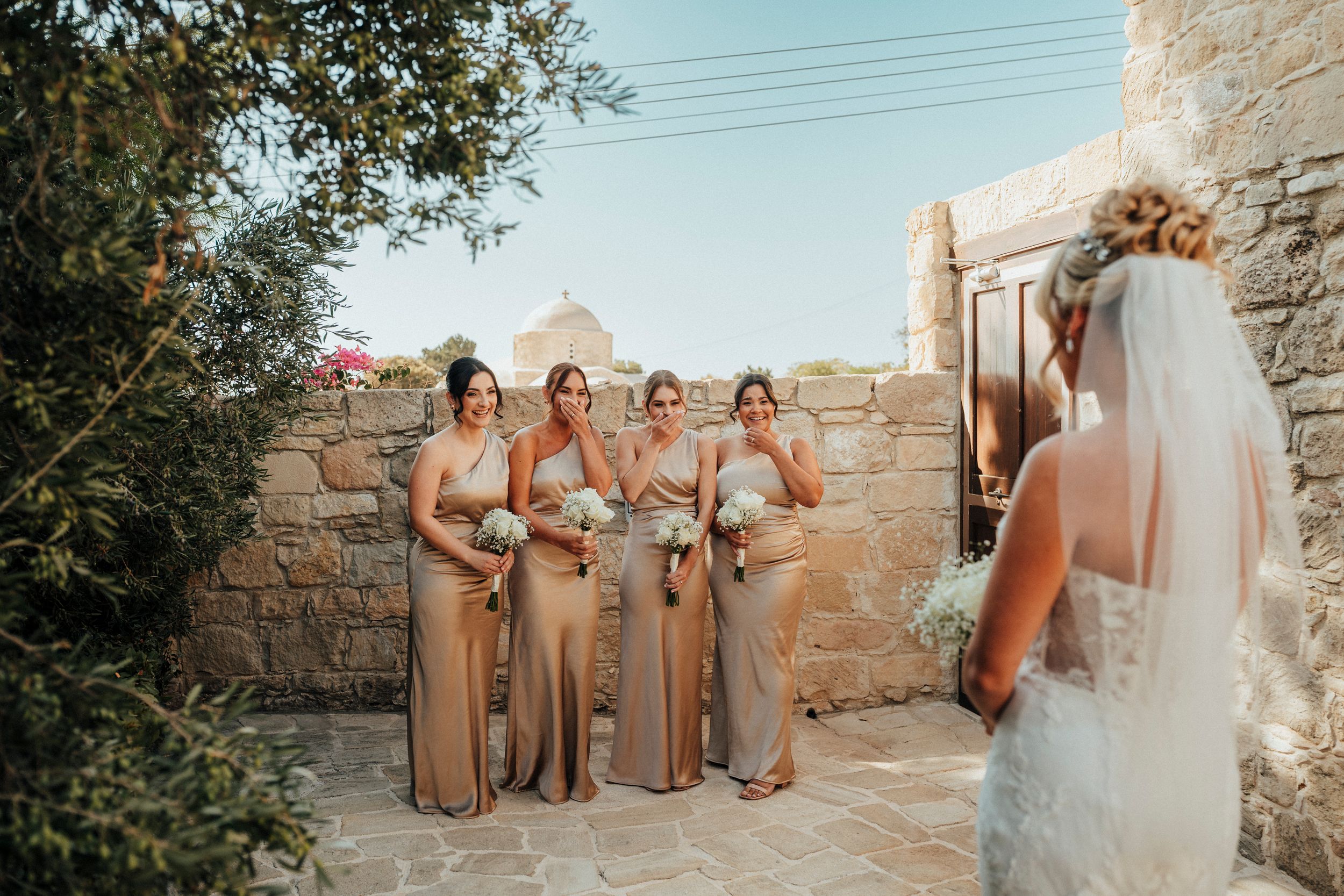 Wedding party in champagne-colored dresses poses together against a rustic stone wall at sunset.