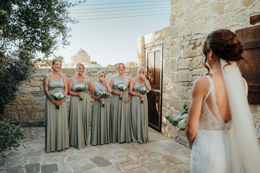 A wedding party in sage green dresses poses together against a rustic stone wall at an outdoor venue.